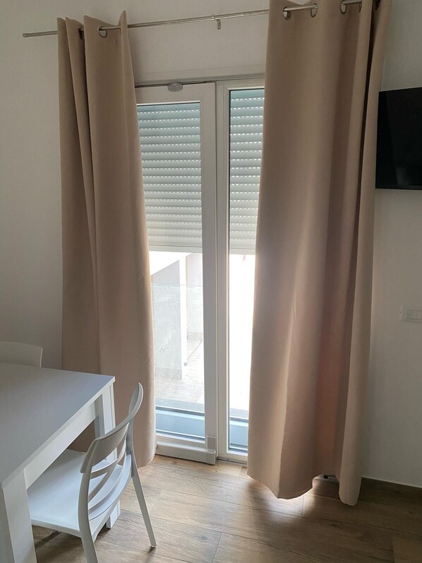A room with a white table and chair, beige curtains, and a sliding glass door leading to an outdoor area.