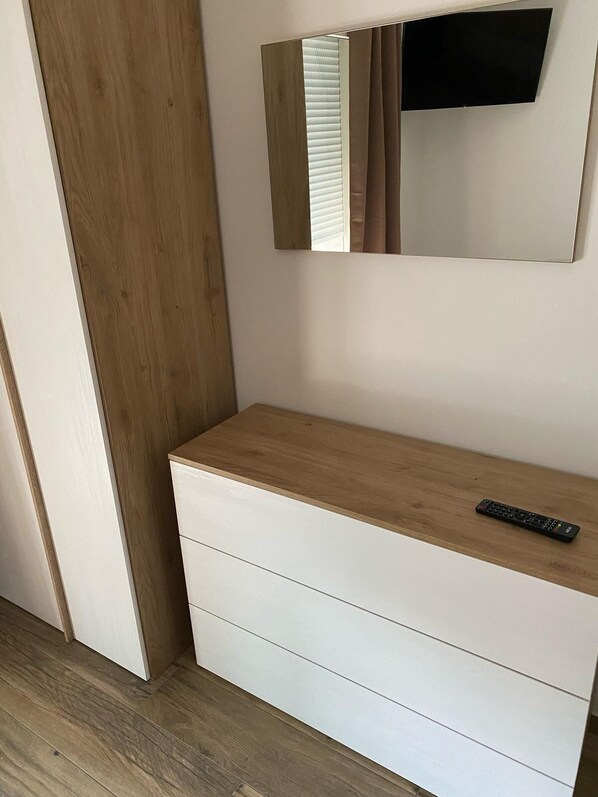 A room with a wooden wardrobe, a mirror, and a remote control on a wooden shelf.