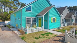 A turquoise house with a white picket fence, a small porch, and a front yard with grass and gravel.