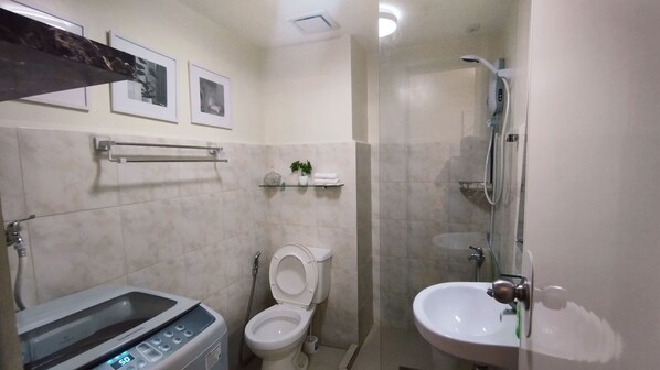 Un baño con inodoro, lavamanos y ducha. Hay cuadros en la pared y una repisa con una planta y toallas.