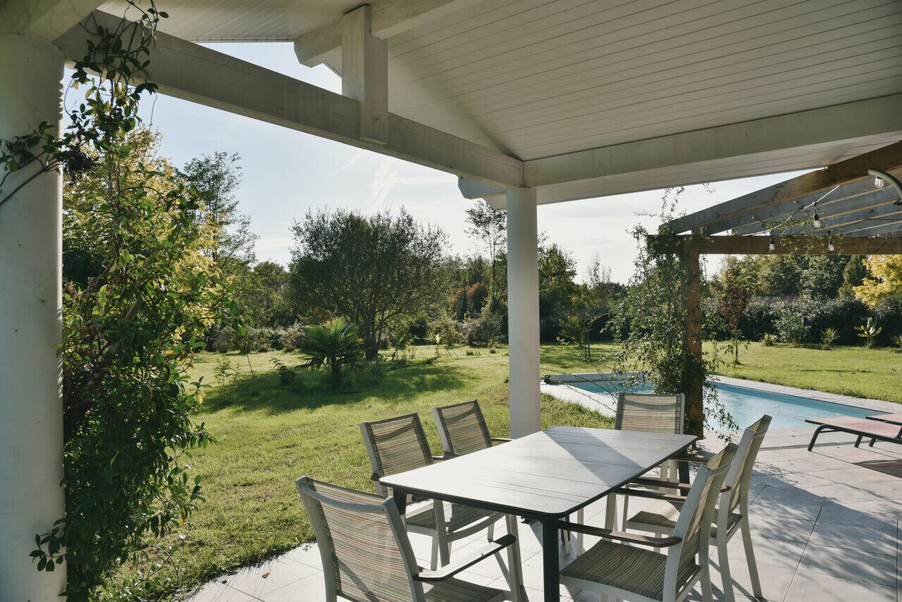 A covered patio with a table and chairs, a pool, and a garden.