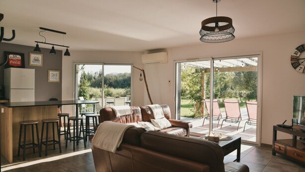 A modern living room with a sofa, a dining table, and a bar area with stools. There is a large sliding glass door leading to an outdoor patio. - l'Acacia, swimming pool and large garden (Bénesse-Maremne)