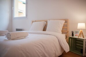 Una cama bien hecha con sábanas blancas y una toalla enrollada, una mesita de noche con una lámpara y un adorno pequeño, y una ventana con vista a un edificio.