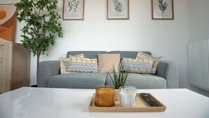 Un salon avec un canapé gris, des coussins à motifs, une petite plante, un plateau avec une bougie et une télécommande, et des reproductions botaniques encadrées accrochées au mur.
