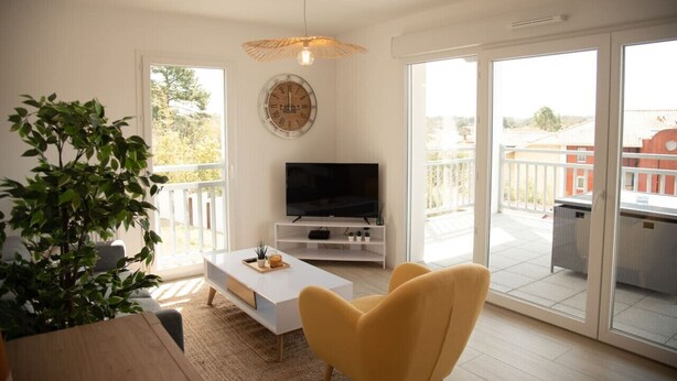 Un salon moderne avec un canapé blanc, un fauteuil jaune, un téléviseur posé sur un meuble blanc, une grande fenêtre et un balcon avec une balustrade.