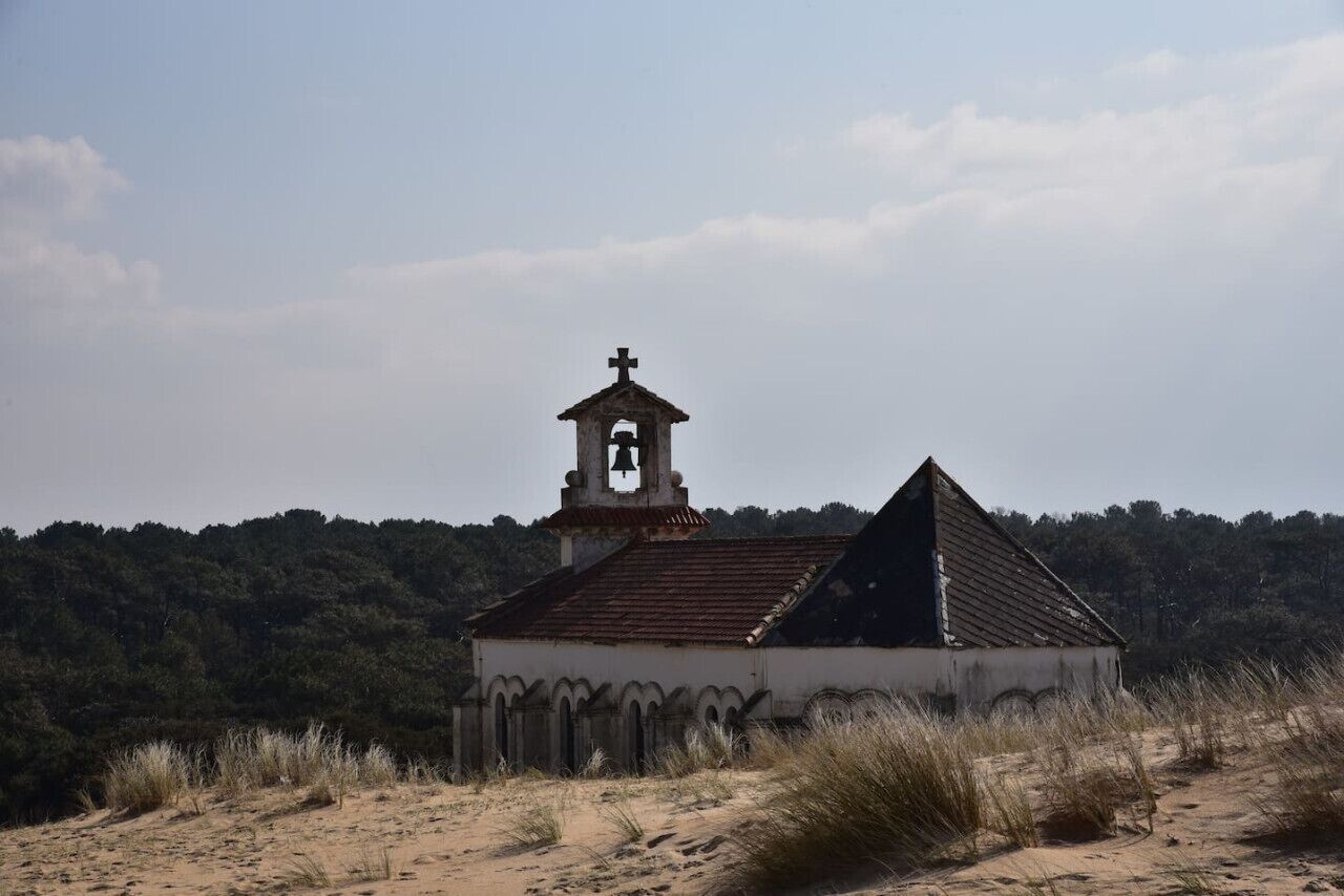 A small, isolated church with a bell tower and a triangular roof, surrounded by dry grass and trees.