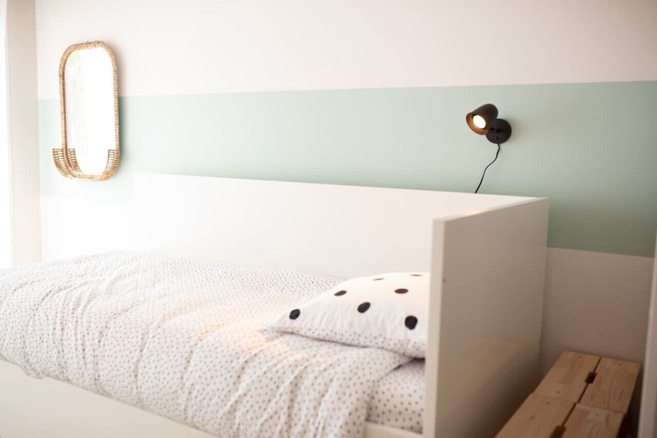 Ein Bett mit weißem Kopfteil, einem gepunkteten Kissen, einer gewebten Wanddekoration und einer an der Wand befestigten Lampe.