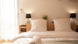 Una cama bien tendida con sábanas blancas y una toalla enrollada, dos mesitas de noche con lámparas, una planta pequeña y una ventana con cortinas translúcidas.
