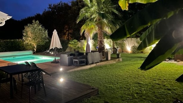 Un patio exterior bien iluminado con piscina, mobiliario y una palmera.