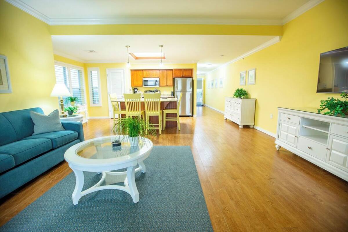 A bright living room with a blue sofa, a glass coffee table, and a kitchen area with wooden cabinets and a refrigerator.