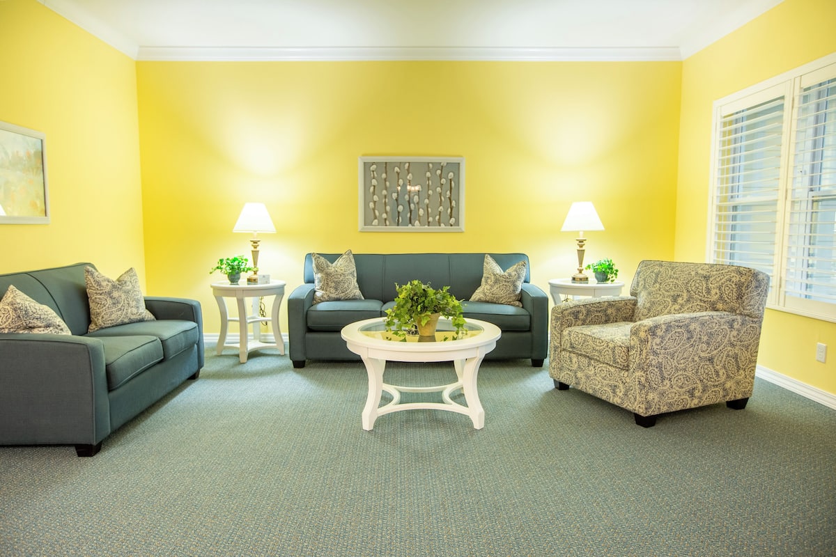 A living room with a yellow wall, a green sofa, a patterned armchair, and a round coffee table with a plant.