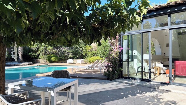 Uma área de piscina com uma sala envidraçada, assentos externos e uma árvore que oferece sombra.