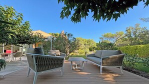 Um pátio externo com mobiliário de vime e uma mesa, rodeado por vegetação e uma casa ao fundo.