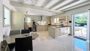 Cozinha espaçosa com ilha central, área de jantar com mesa de mármore e vista para o exterior através de grandes janelas.