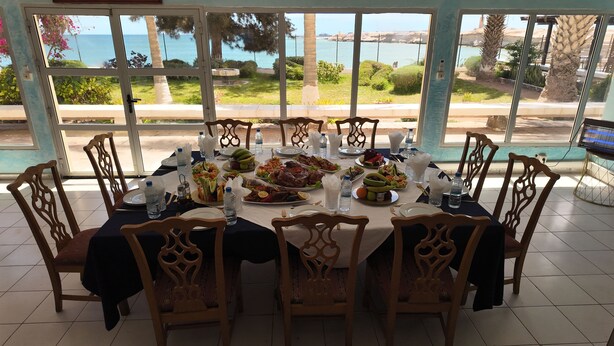 Une table à manger, dressée pour huit personnes, entourée de chaises et bordée de grandes fenêtres offrant une vue sur la mer et la verdure.