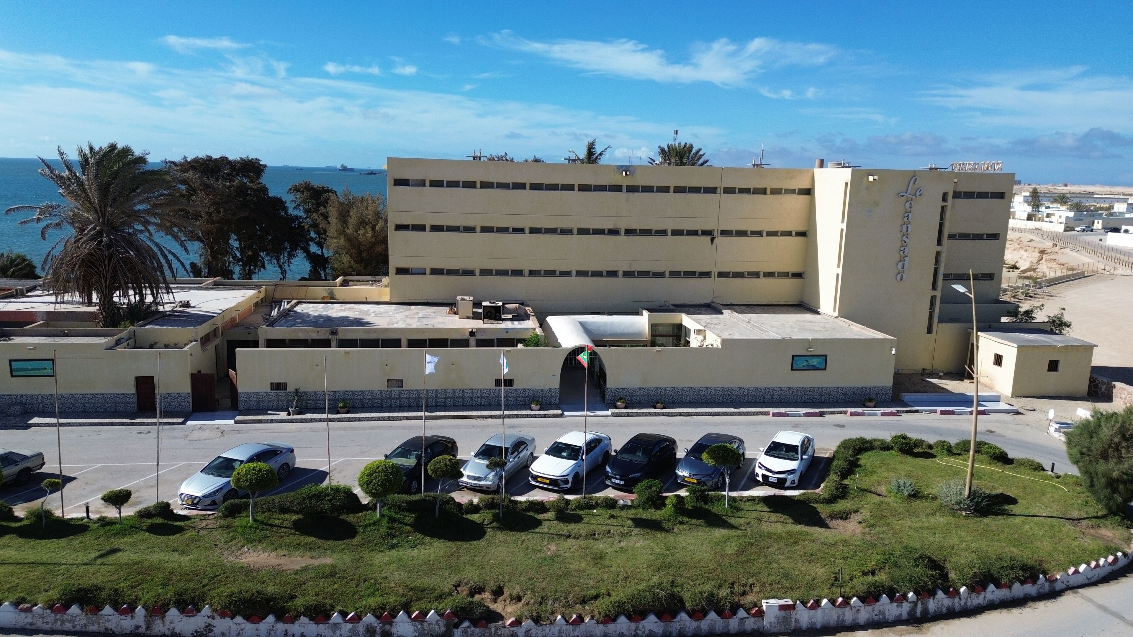 A multi-story building with a parking lot in front, a flag, and a view of the sea.