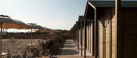 Une rangée de cabanes en bois, situées sur la plage, est reliée à une zone de plage sablonneuse par une passerelle en bois.