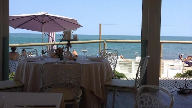 A dining area with tables and chairs, overlooking a beach with people and umbrellas.