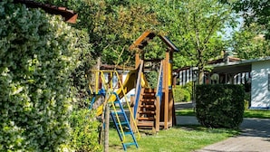 A wooden play structure with slides and a ladder in a garden area with potted plants and a paved walkway.