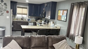 Ein modernes Wohnzimmer mit einem braunen Ledersofa, einer Küchenzeile mit dunklen Schränken und einem Deckenventilator.