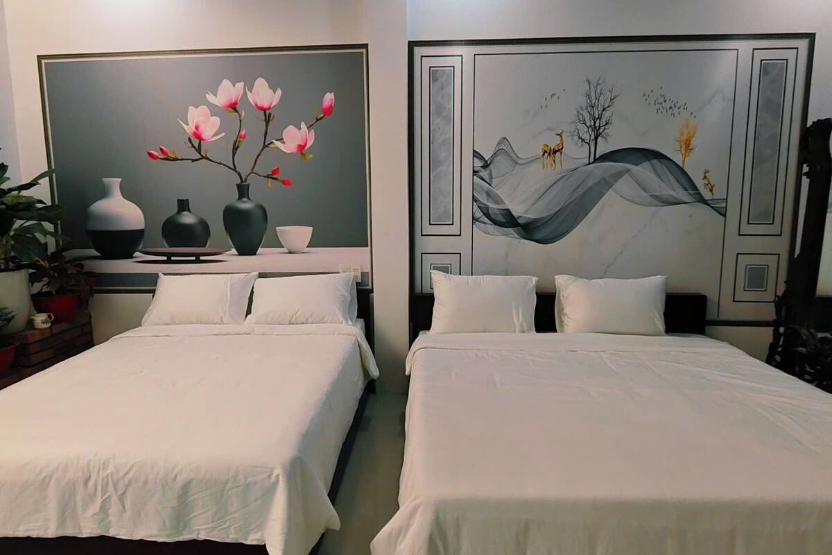 A hotel room with two beds, a large painting on the wall, a mirror, and several potted plants.