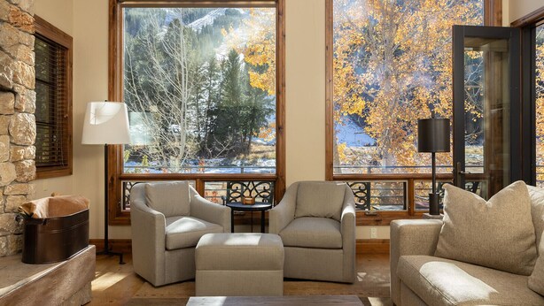 A cozy living room with a stone wall, large windows with a view of autumn trees, a fireplace, and comfortable seating. - Warm Springs 4-bedroom, 4 bath. Gourmet kitchen, fitness room, pool, and hot tub (Ketchum)