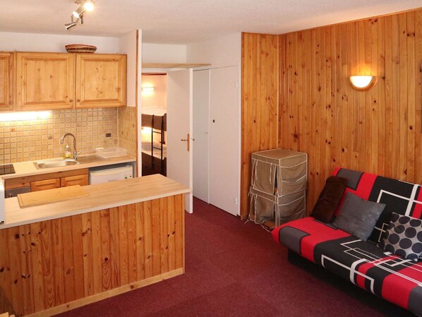 A compact kitchen with wooden cabinets and a built-in sink, a small dining area with a bench, and a red carpeted living space with a sofa and cushions.