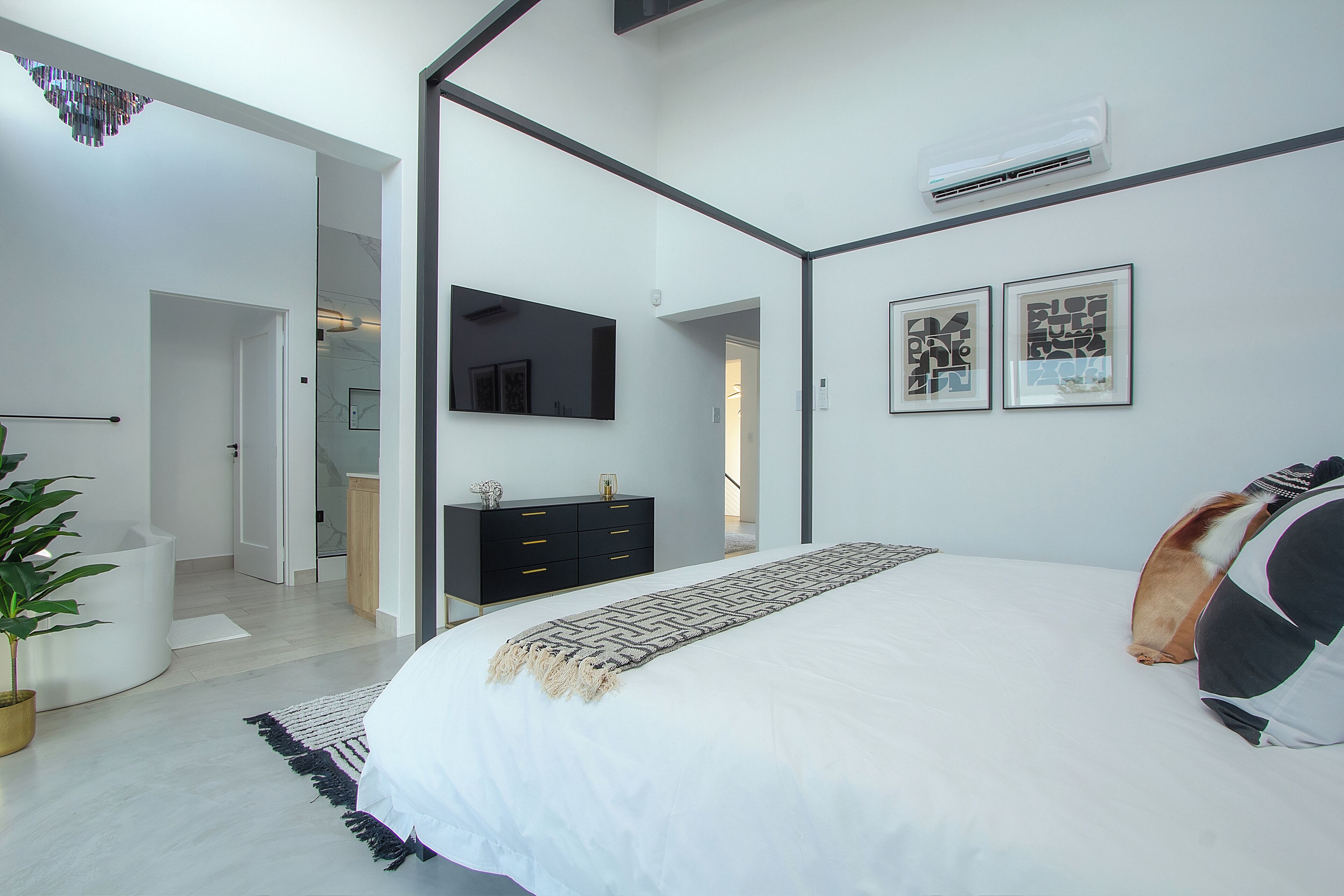 Une chambre moderne avec un grand lit, une commode noire, un téléviseur à écran plat et une plante en pot.