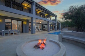 A modern house with a fire pit in the center, outdoor seating, and a pool in the background.