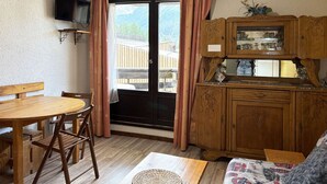 A cozy room with a wooden dining table, chairs, a woven mat, a sofa, a TV, and a wooden cabinet with a view of mountains through the window.