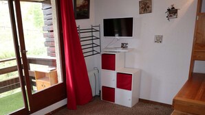 Ein Zimmer mit Fernseher, rotem Vorhang, Holztisch und weißem Schrank mit roten Schubladen.