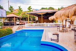 A swimming pool with a blue border, a covered seating area with a thatched roof, and a fenced patio with tables and chairs.