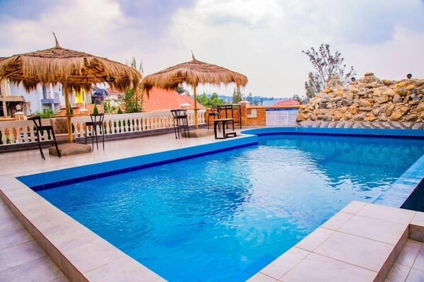 A swimming pool with a tiled deck, two thatched umbrellas, and a stone wall.