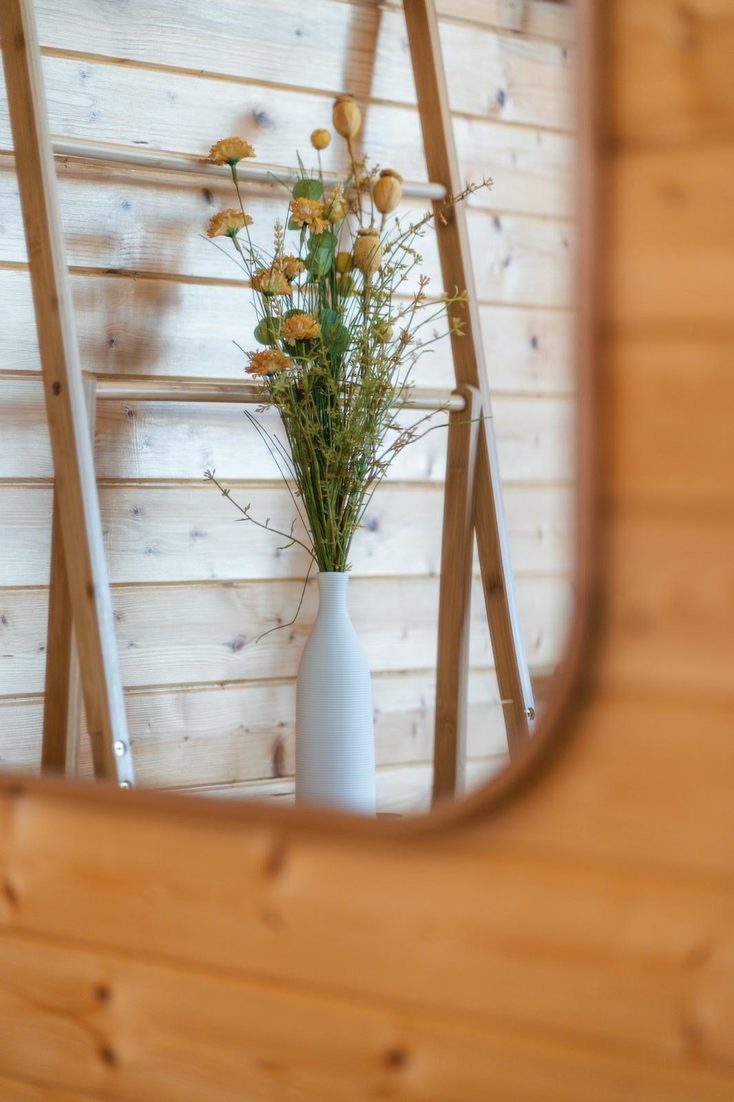 A white vase with dried flowers and greenery on a wooden wall.