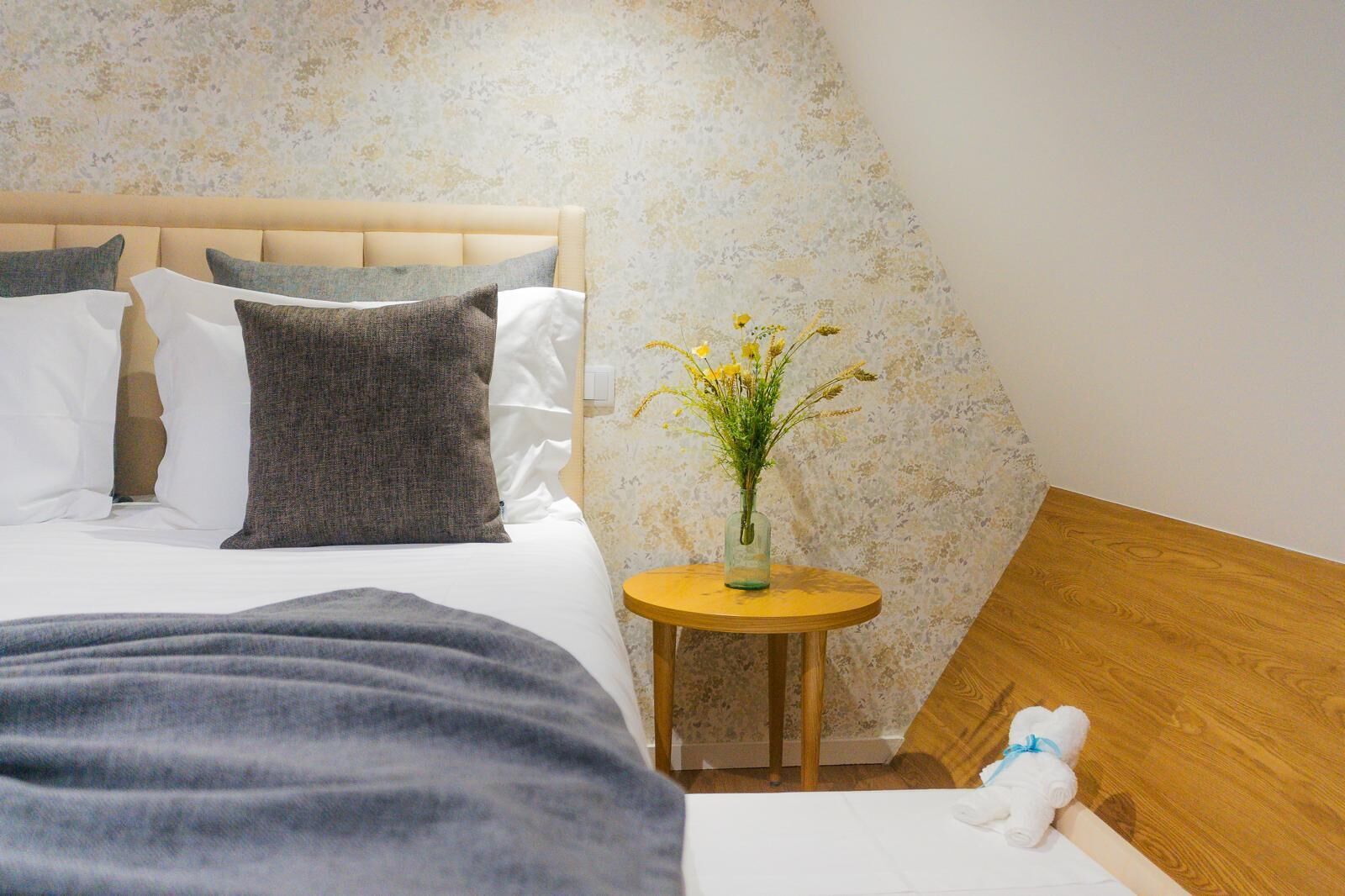 Un dormitorio con cama, almohadas, un jarrón con flores, una mesita redonda pequeña y una mesita de noche de madera.