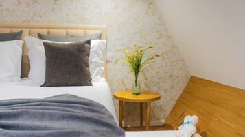 Un dormitorio con cama, almohadas, un jarrón con flores, una mesita redonda pequeña y una mesita de noche de madera.