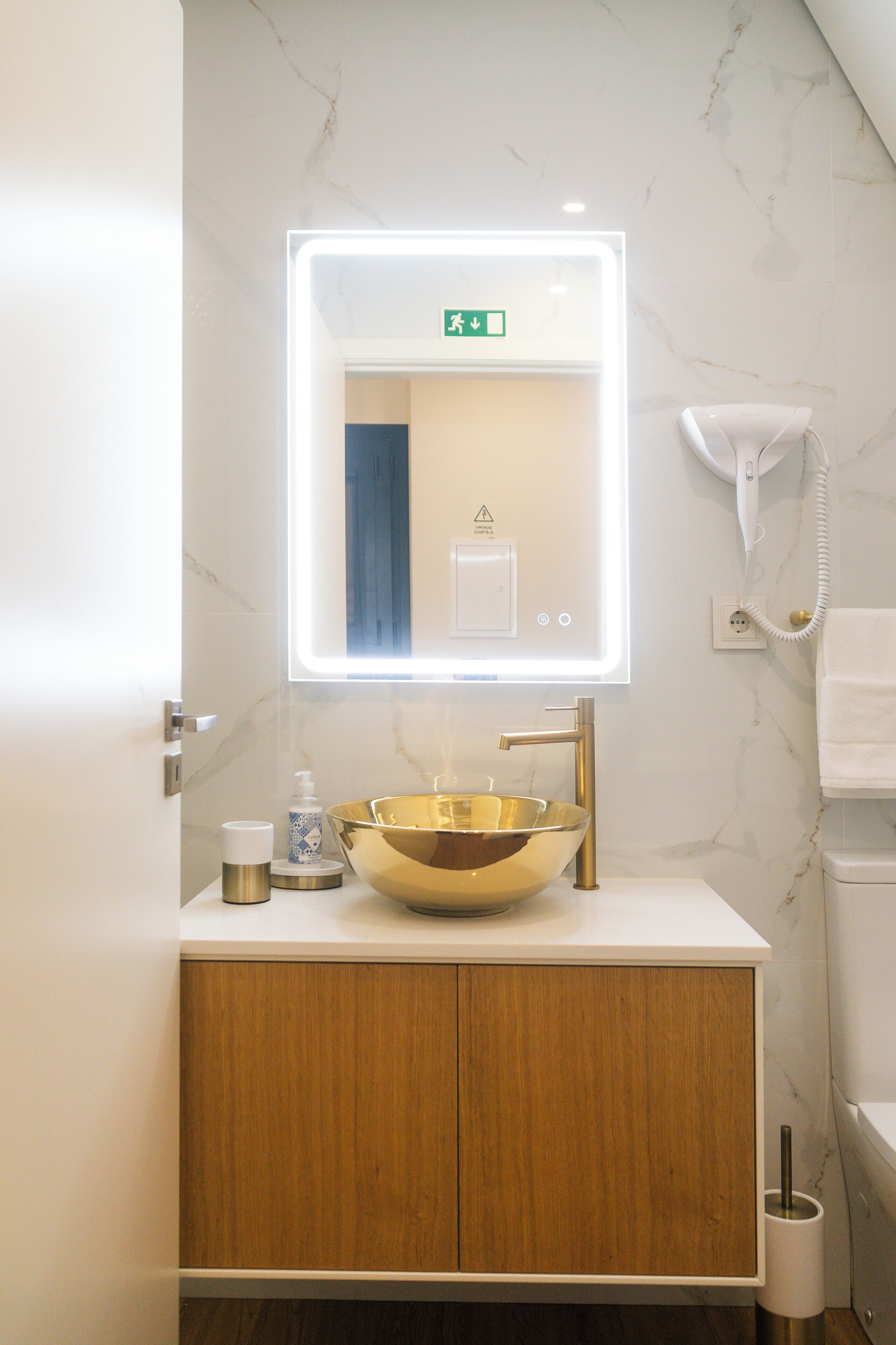 A bathroom with a marble wall, a gold sink, a wooden cabinet, a mirror with a light, and a hairdryer on the wall.