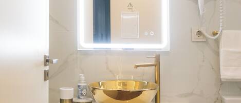 Une salle de bain avec un mur en marbre, un lavabo doré, un meuble en bois, un miroir avec un éclairage intégré et un sèche-cheveux fixé au mur.