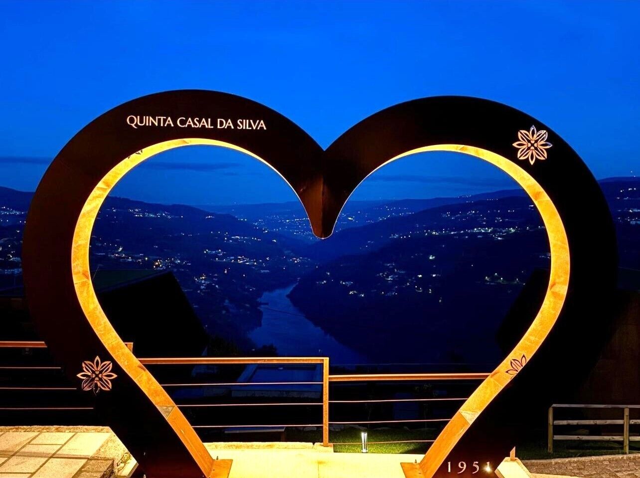 Un panneau en forme de cœur portant l’inscription « QUINTA CASAL DA SILVA » et représentant une vue sur une ville illuminée la nuit.