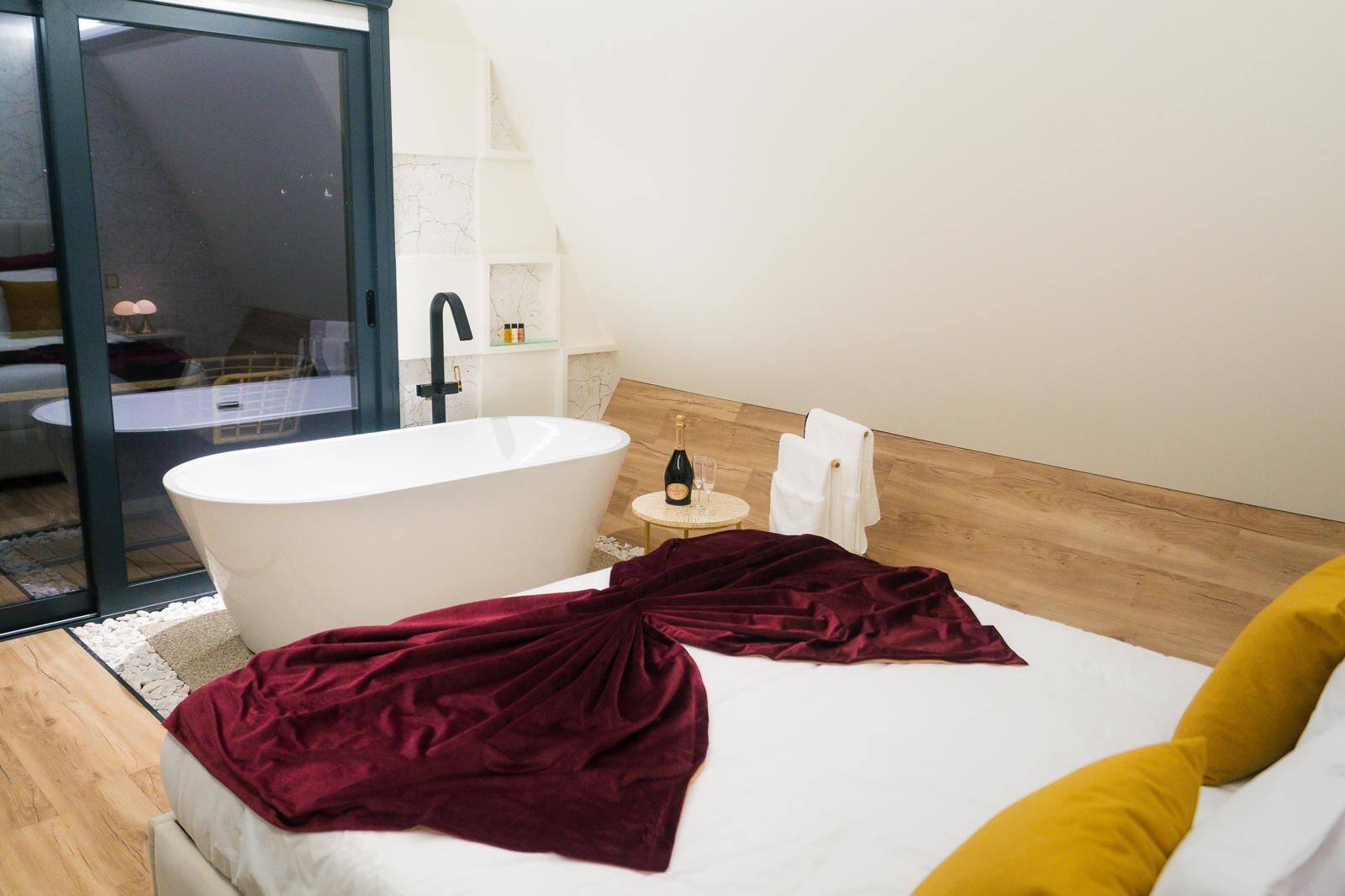 A modern bathroom with a freestanding bathtub, a small round table with a bottle and glass, and a bed with a red blanket and yellow pillow.