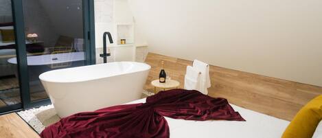 A modern bathroom with a freestanding bathtub, a small round table with a bottle and glass, and a bed with a red blanket and yellow pillow.