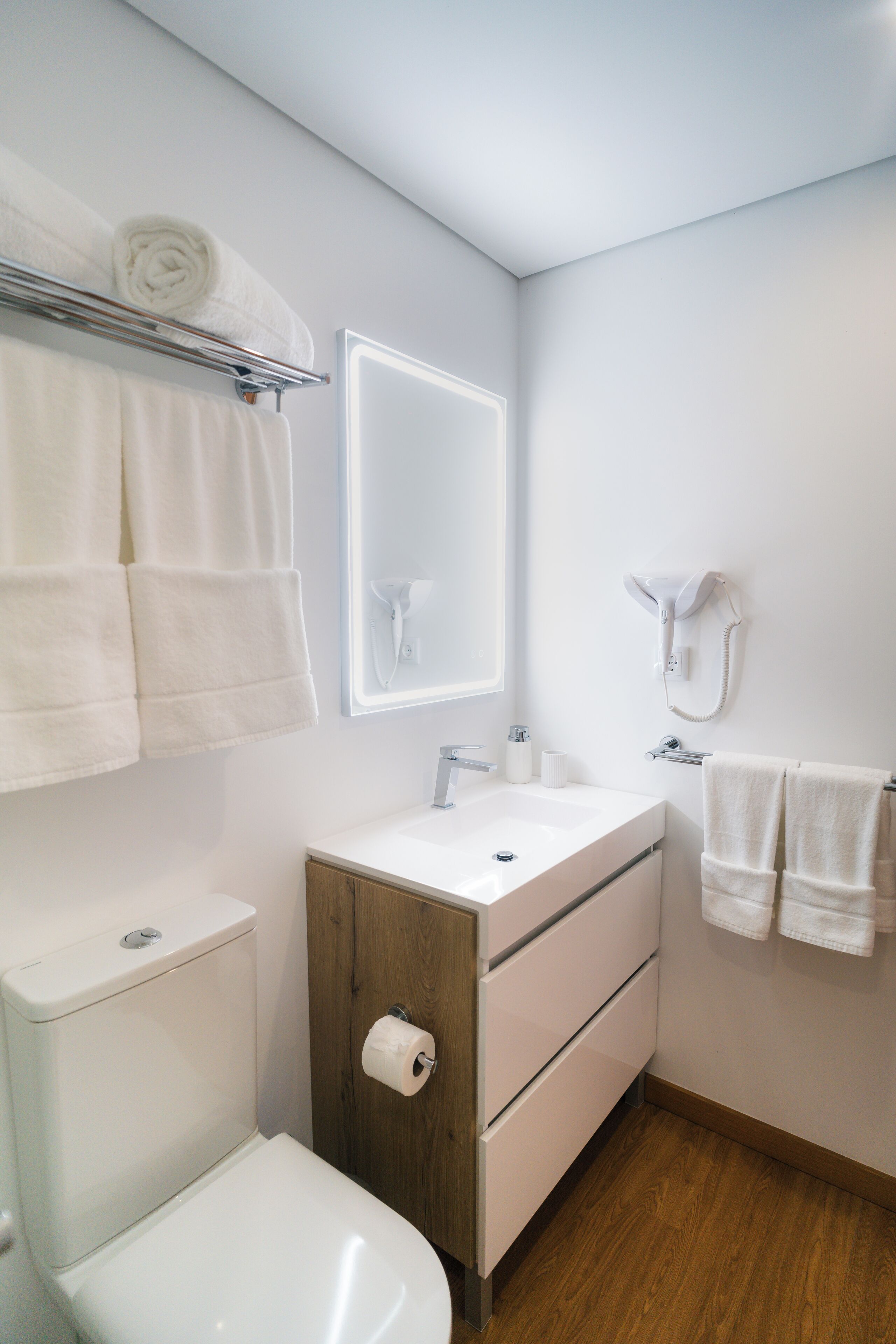 Un baño moderno con inodoro, lavabo y espejo. Hay toallas en un perchero y un secador de pelo en la pared.