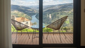 A bedroom with a large sliding glass door leading to an outdoor deck with two wicker chairs and a table, overlooking a river and hilly landscape. - Quinta Casal da Silva 1951 (Marco de Canaveses)