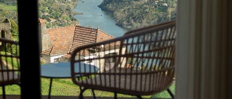 Un balcón con una silla de mimbre y una mesa, con vistas a un valle con río y colinas lejanas.