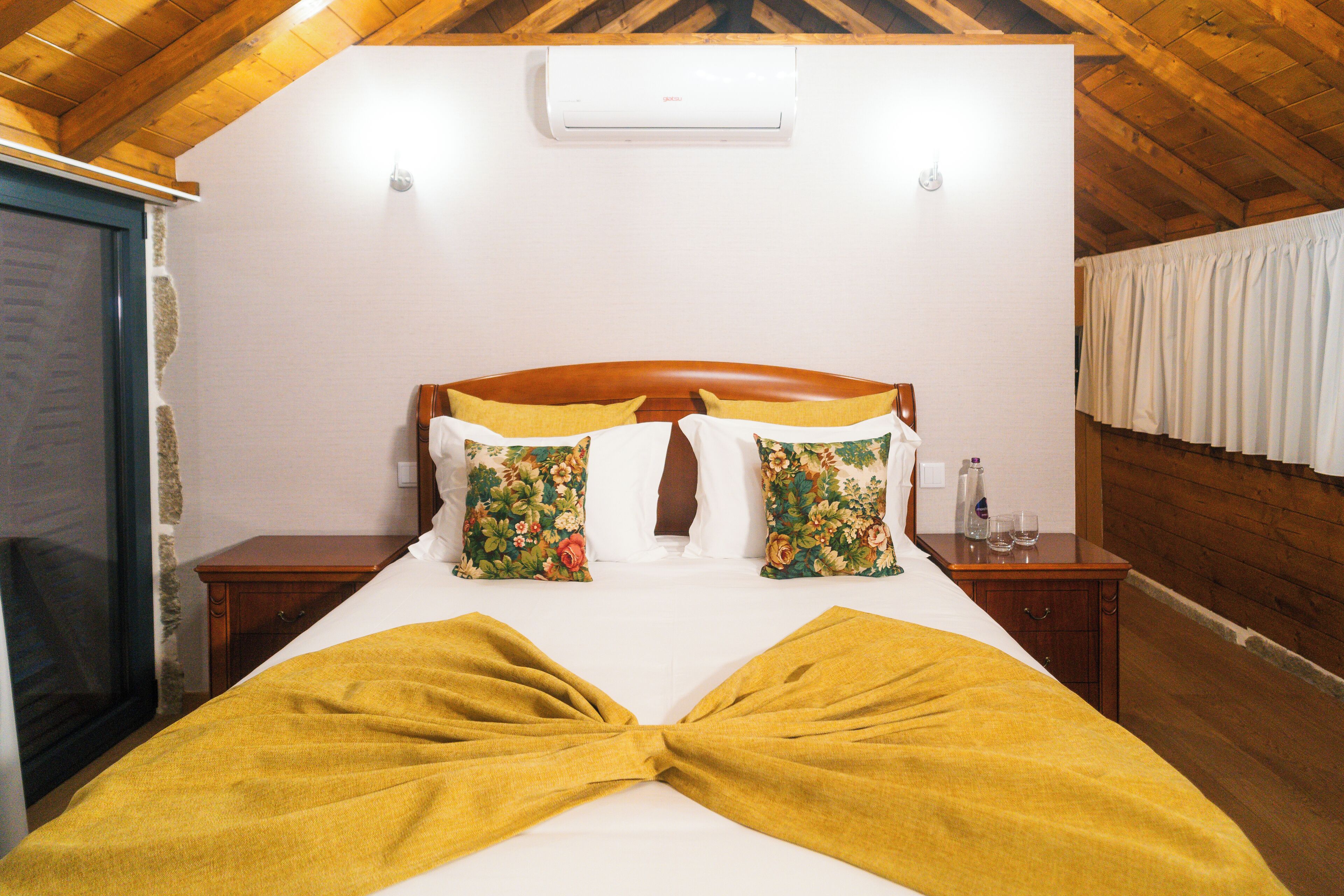 A bedroom with a wooden ceiling, a bed with floral pillows, a yellow throw blanket, and a nightstand with a bottle and glasses.