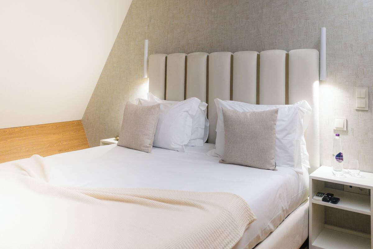 Ein modernes Hotelzimmer mit einem großen Bett, einem Kopfteil mit vertikalen Paneelen, Nachttischen mit Lampen und einem Beistelltisch mit Flasche und Gläsern.