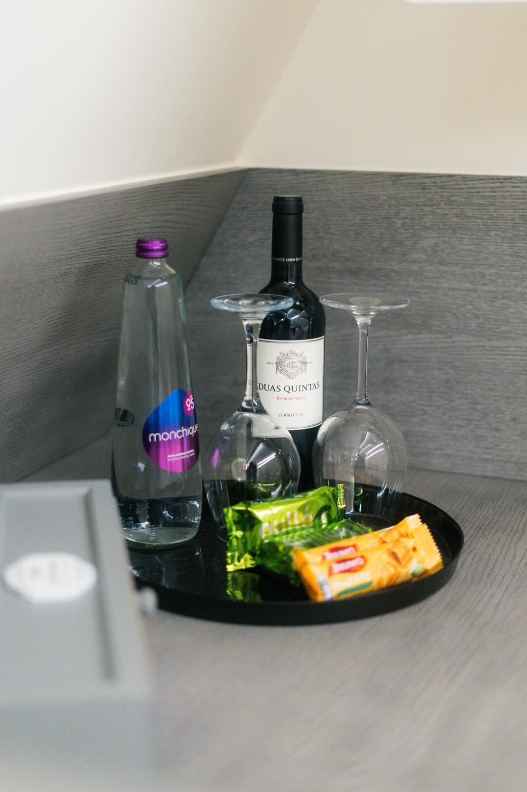 A minibar setup with a bottle of Duas Quintas wine, a bottle of monchiquo, two glasses, and a snack package.