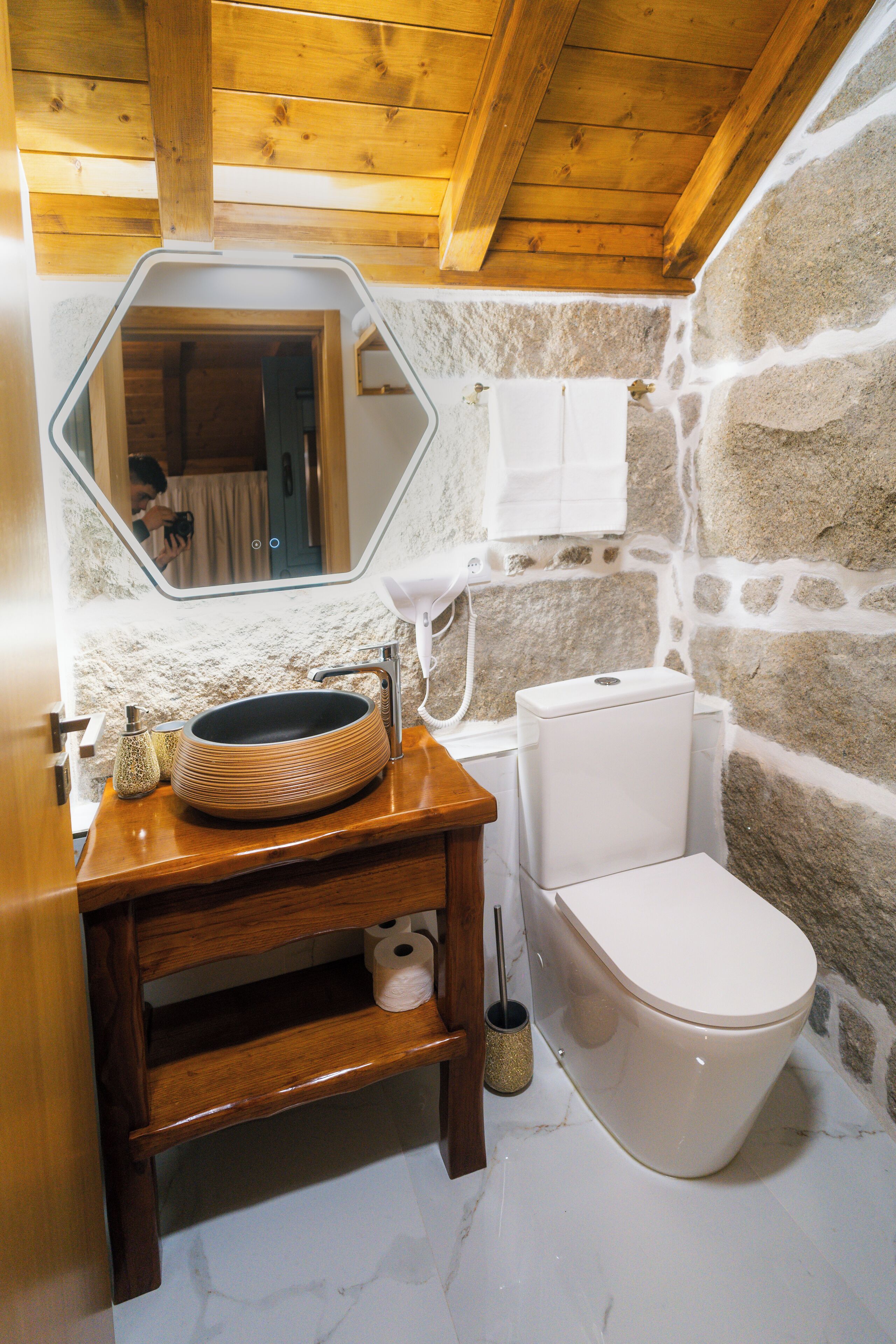 Une salle de bain avec un meuble-lavabo en bois, un lavabo rond, des toilettes et un mur en pierre.