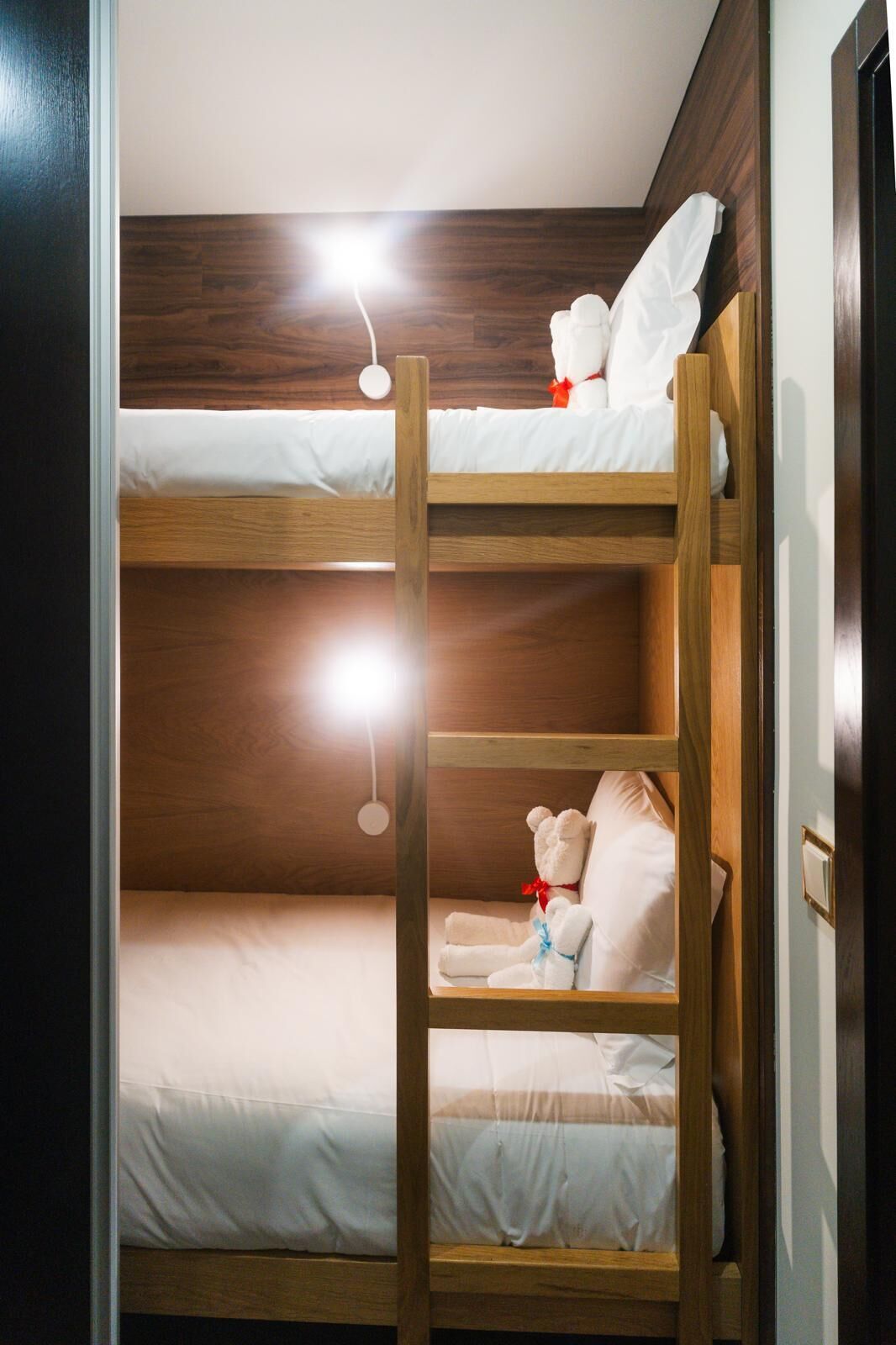 Una cama litera con dos niveles, cada uno con un colchón blanco y estructura de madera. Un pequeño oso de peluche blanco está sobre la cama superior.