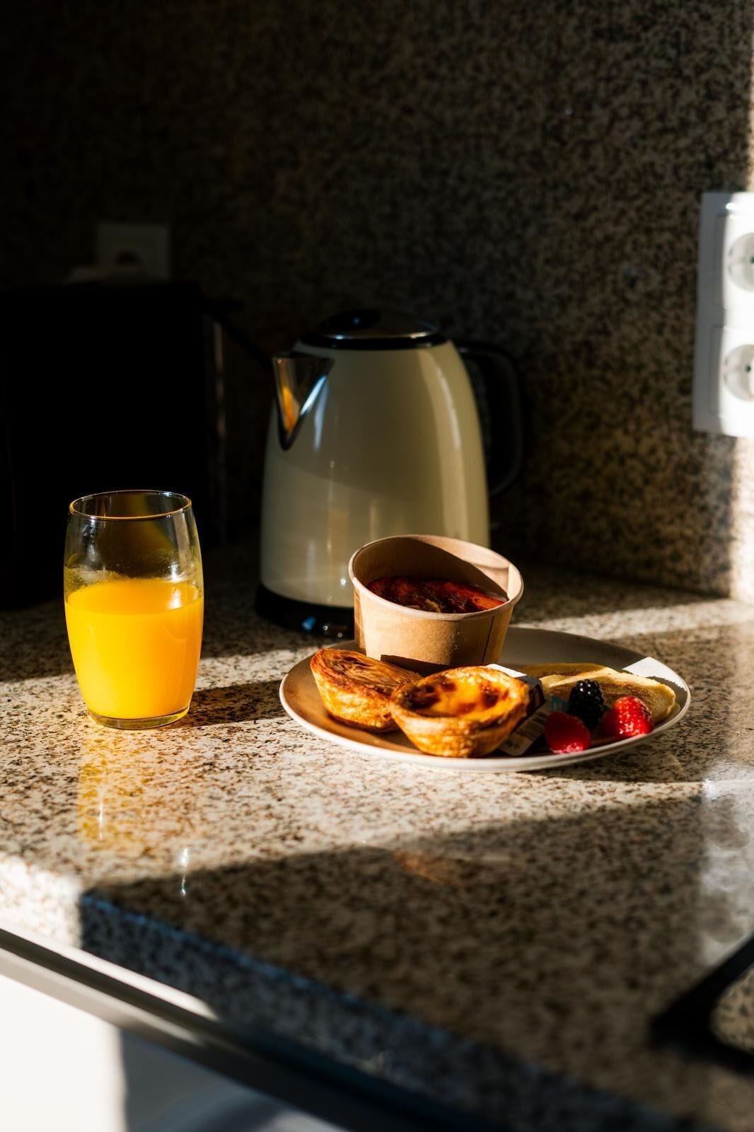 Une table dressée pour le petit-déjeuner, avec un verre de jus d’orange, une tasse de thé et une assiette contenant des viennoiseries et des fruits rouges, le tout posé sur un plan de travail en granit.